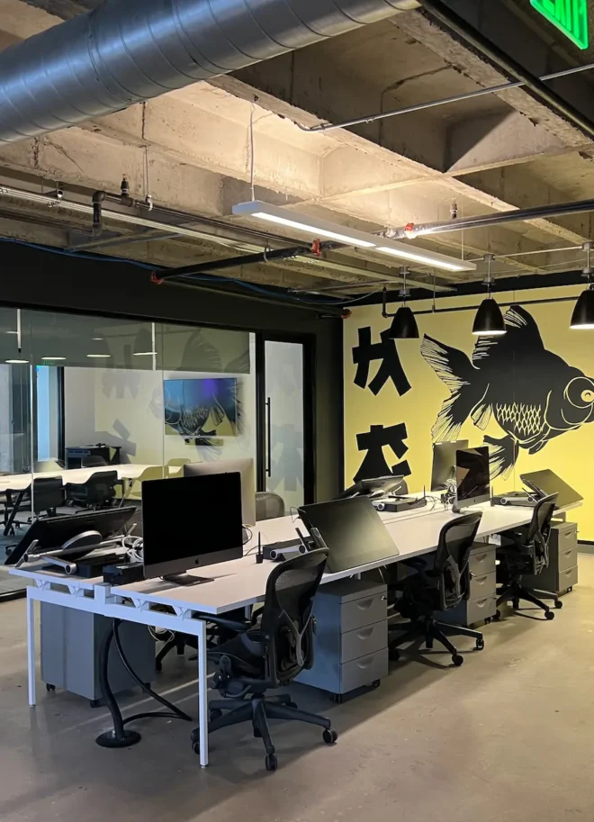 Open office workspace with desks, monitors, and a large fish mural next to a glass-walled conference room