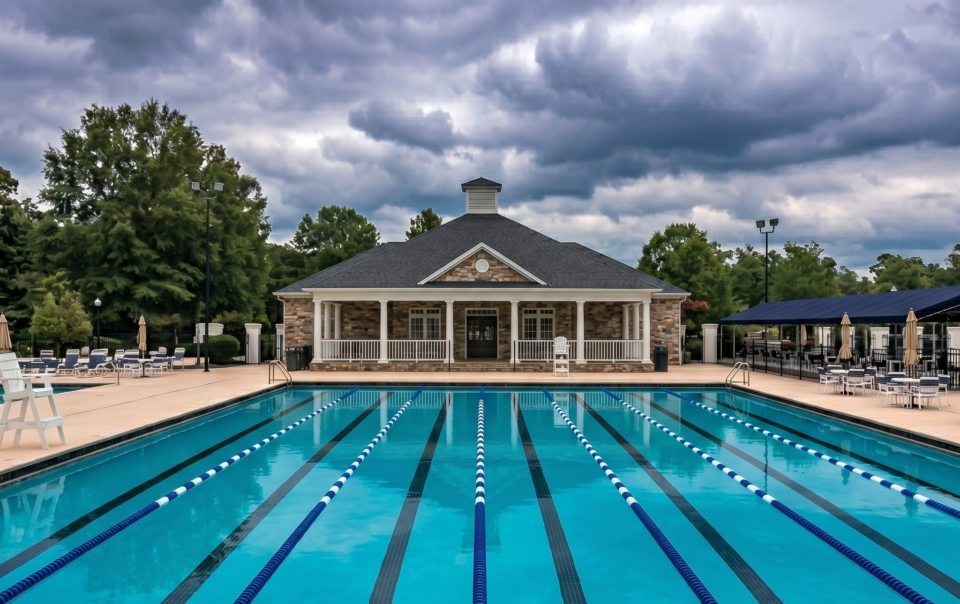 Symmetrical low-angle photograph of a multi-lane competitive swimming pool with blue and white lane ropes, leading the eye to a stone facade clubhouse under a dramatic, stormy cloud-filled sky. A lifeguard stand is visible on the left.